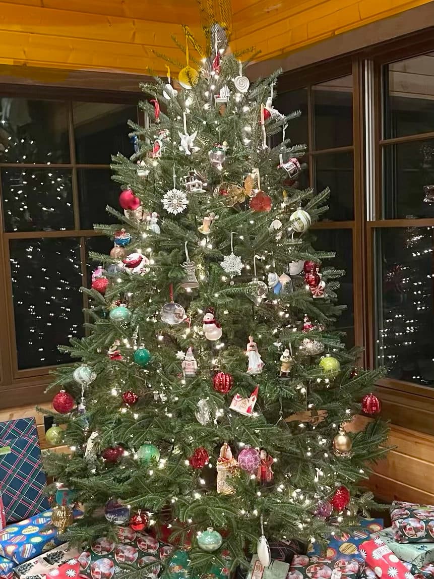 A Fraser Fir from our farm decorated with lights and glass ornaments in a wood-paneled farmhouse room, with wrapped presents underneath
